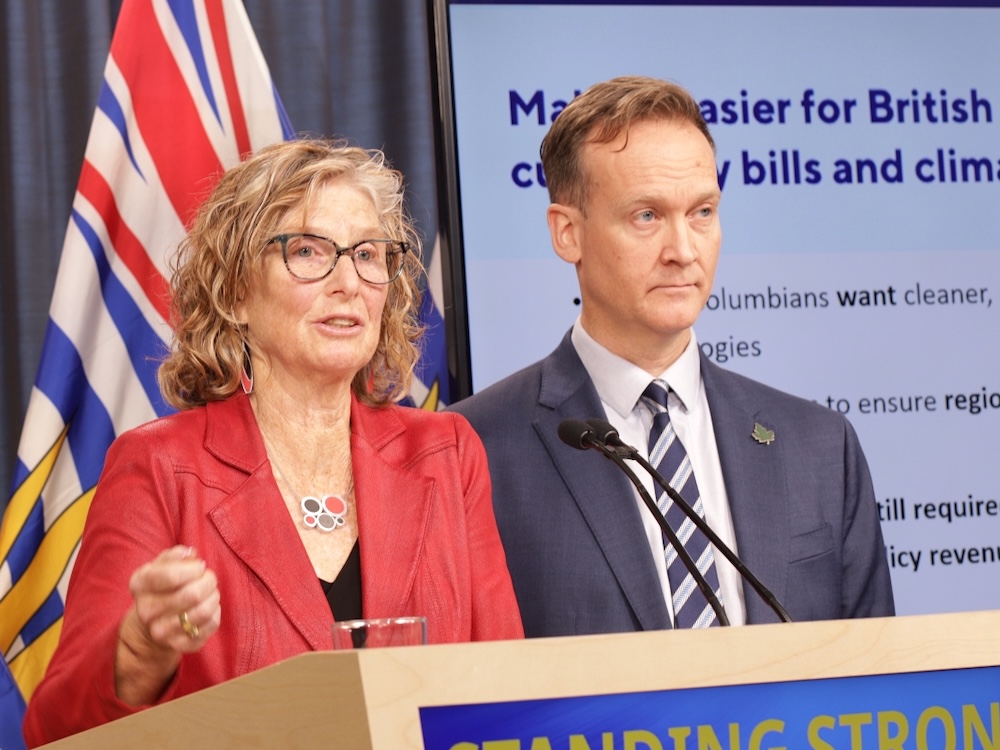 Merran Smith is speaking in front of a microphone. She has blond hair and glasses and is wearing a red blazer. Dan Woynillowicz stands beside her. He is slightly taller and is wearing a blue suit.