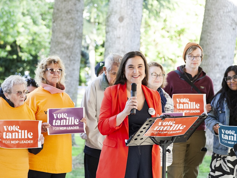 A woman with long brown hair wearing a red blazer holds a microphone and smiles at the camera. Several people hold signs that read &lsquo;Vote Tanille for NDP leader.&rsquo;