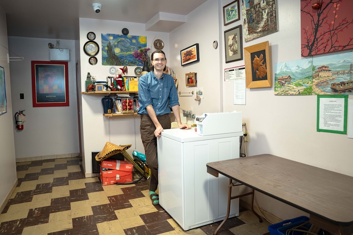 A man with light skin and brown hair stands in the laundry room of an apartment building. There are a number of prints and paintings on the walls. 