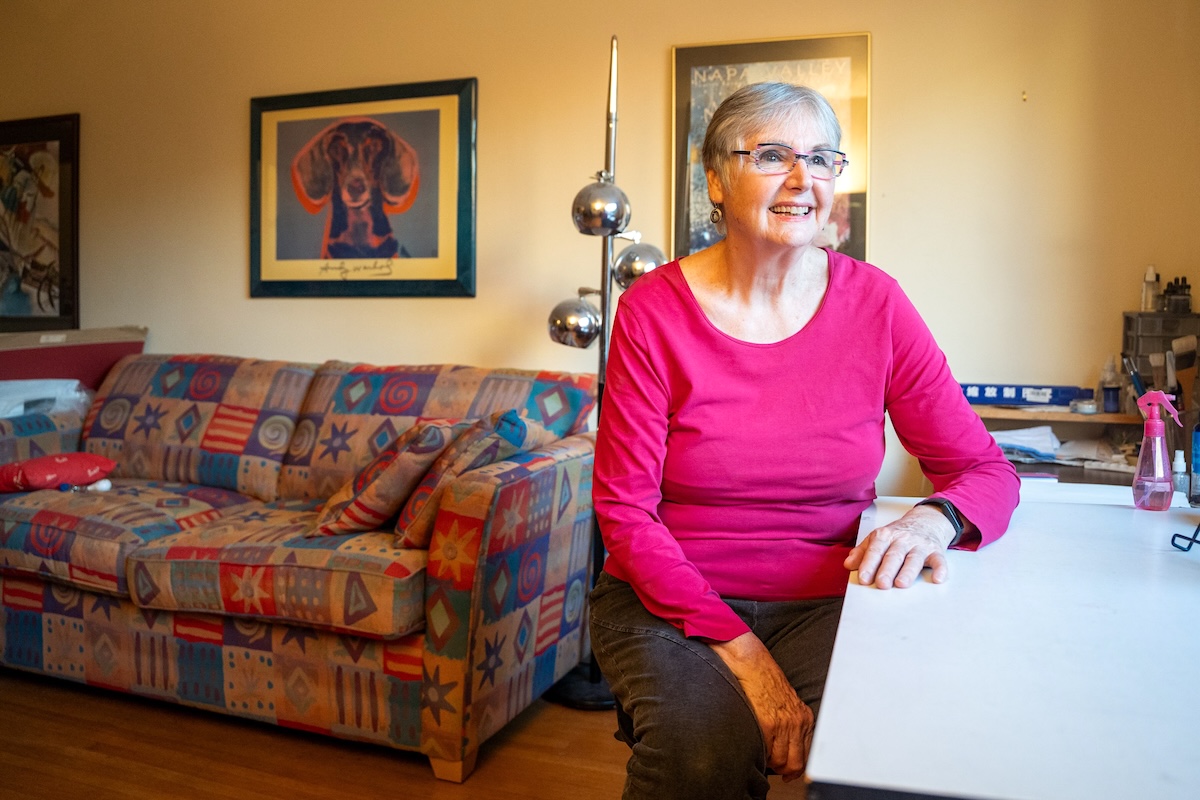 A woman in her 70s with short grey hair sits at a desk in an apartment. A brightly coloured couch and art on the walls can be seen in the background.