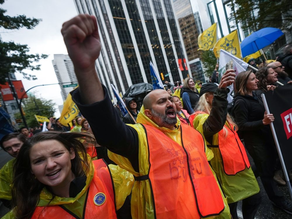 A man wearing a bright orange vest pumps his fist in the air. He is surrounded by a crowd of other people, many of whom are waving yellow flags, the symbol of the BC General Employees&rsquo; Union.