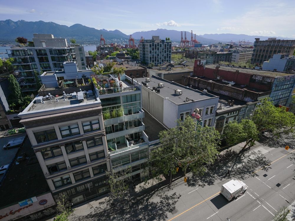 An aerial view of several condo and single-room occupancy hotel buildings in Vancouver&rsquo;s Downtown Eastside neighbourhood. The North Shore mountains and several port cranes can be seen in the distance.