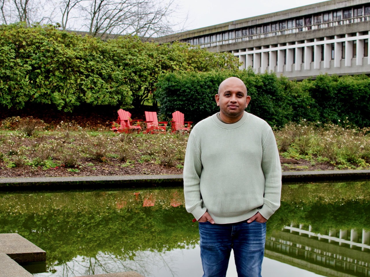 A bald man wearing blue jeans and a white sweater stands in front of a fountain and garden at Simon Fraser University.