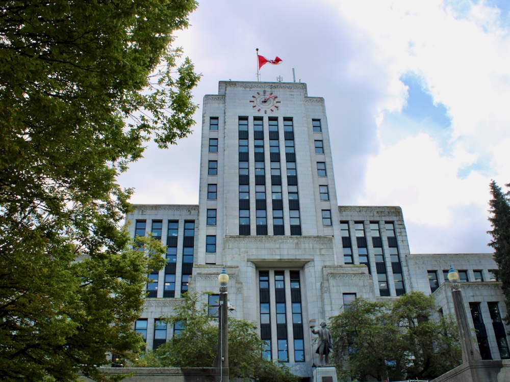 Vancouver City Hall, a grey stone art deco building with a neon clock and a Canadian flag flying from a rooftop flagpole.