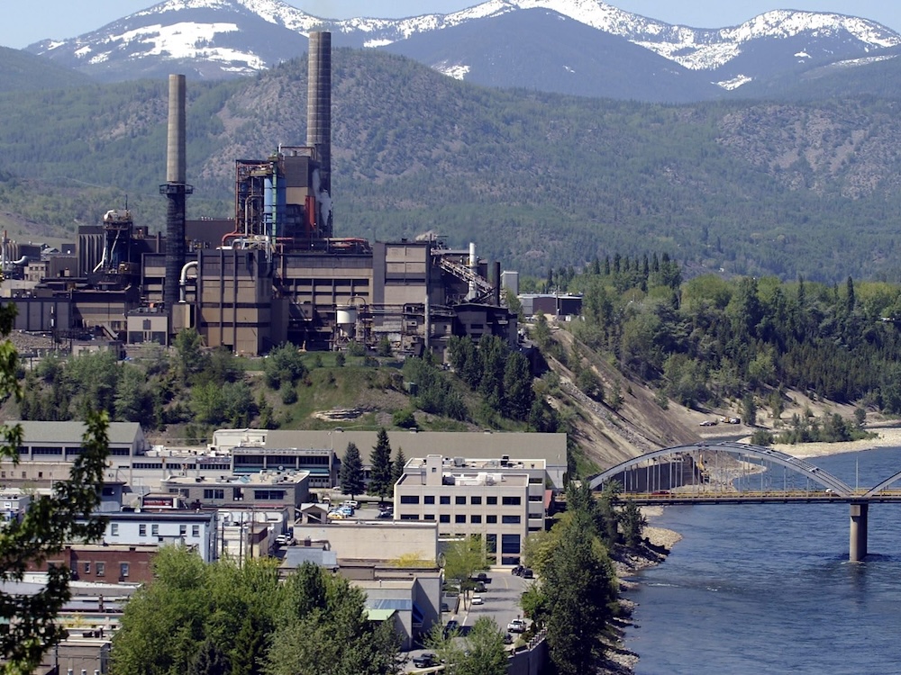A major factory with smokestacks sits beside a river. A bridge spans the river.