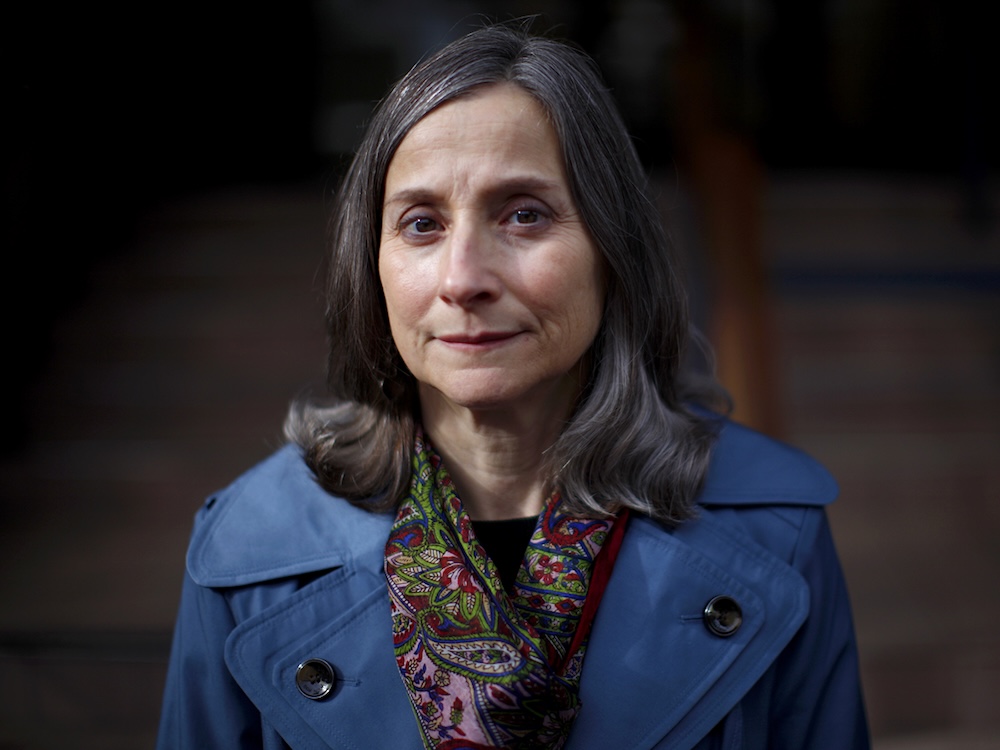 Lisa Lapointe, a middle-aged woman with light skin tone and greying brown hair to her shoulders, poses outdoors for a portrait in gloomy lighting.