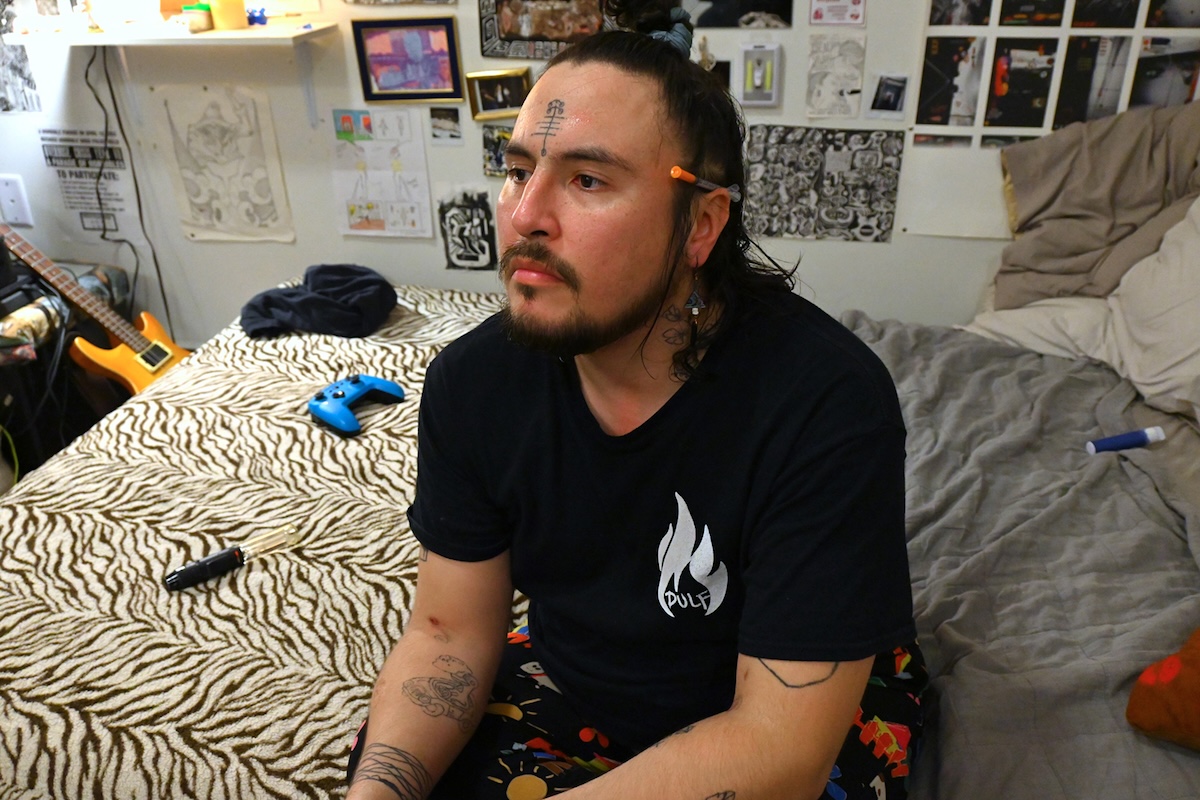 TJ Felix, a non-binary person of Secwe̓pemc descent, has medium-light skin tone, dark hair and a forehead tattoo. They have a capped needle tucked behind their ear and are sitting on their bed in their room. 