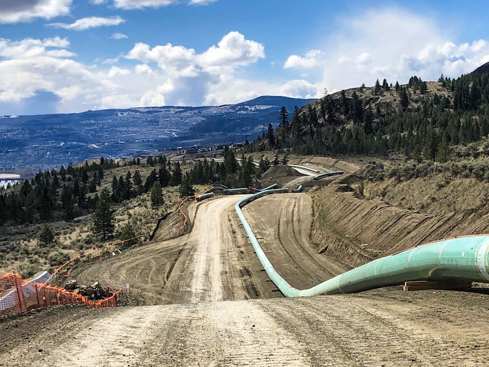 A green pipeline recedes into the distance through a rugged hillside above a river.