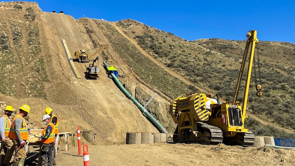 Crews using heavy machinery work to install a pipeline up a small hill in a rugged, arid landscape on a clear day.