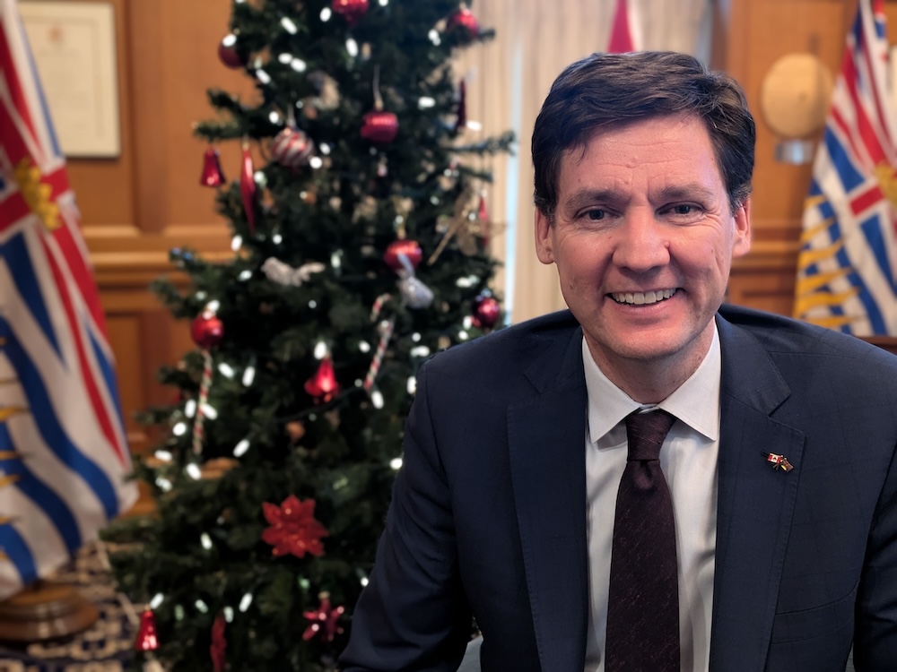 David Eby, a 49-year-old light-skinned man with dark hair, smiles at the camera. He wears a blue suit and sits in an office with a Christmas tree.