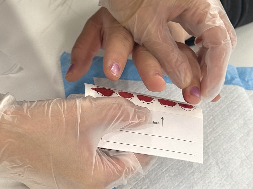 A woman holds another person&rsquo;s hand. Their finger has been pricked. It is being placed against a card to leave a blood spot.