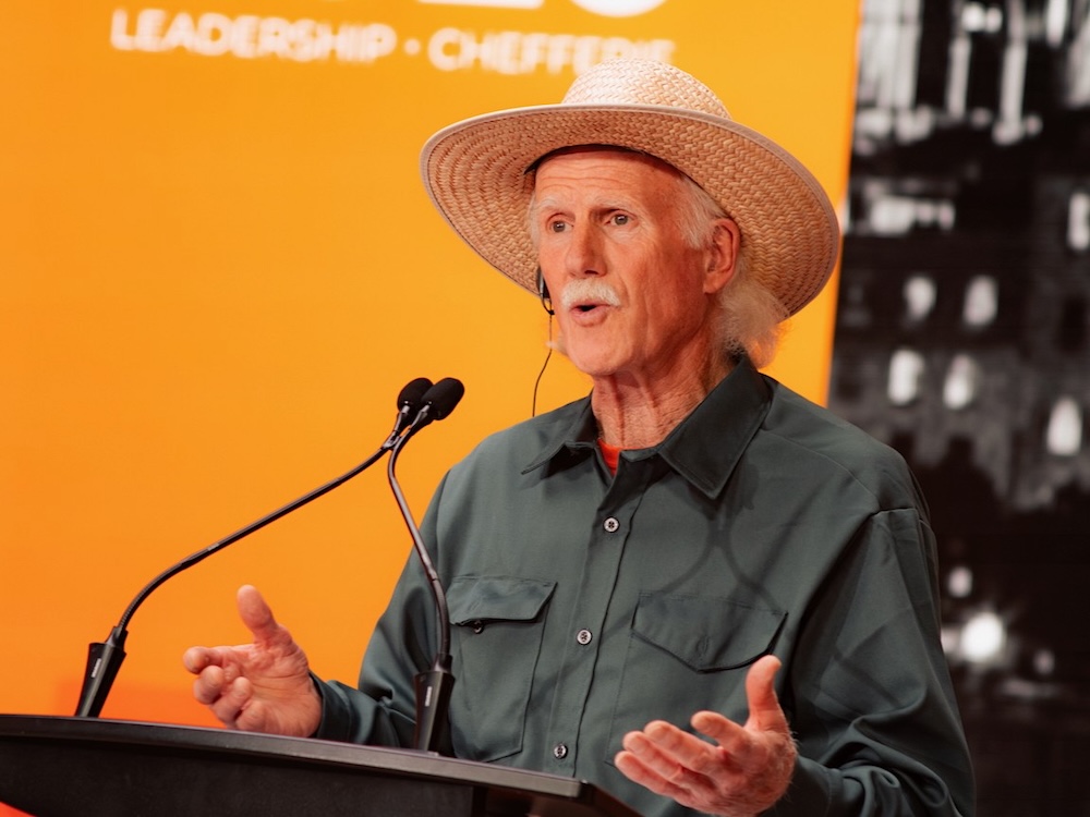 A man in his 70s with white hair and wearing a straw, wide-brimmed hat speaks at a podium. He wears a western-style button-down shirt.