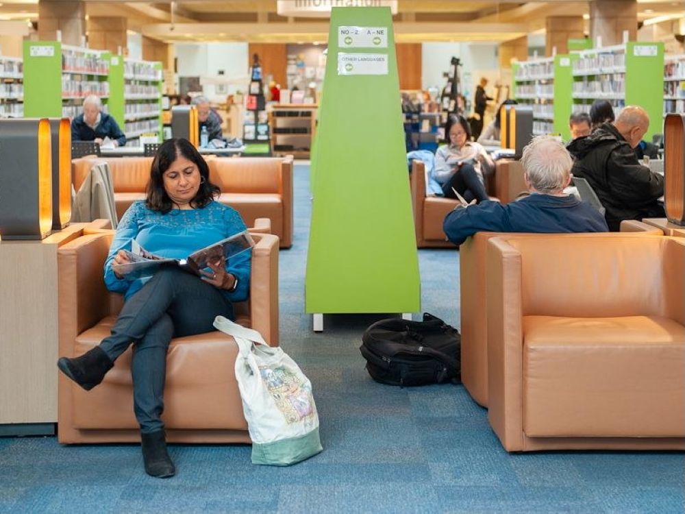 A woman wearing a blue sweater sits in an armchair reading a book inside a bright, modern public library building.
