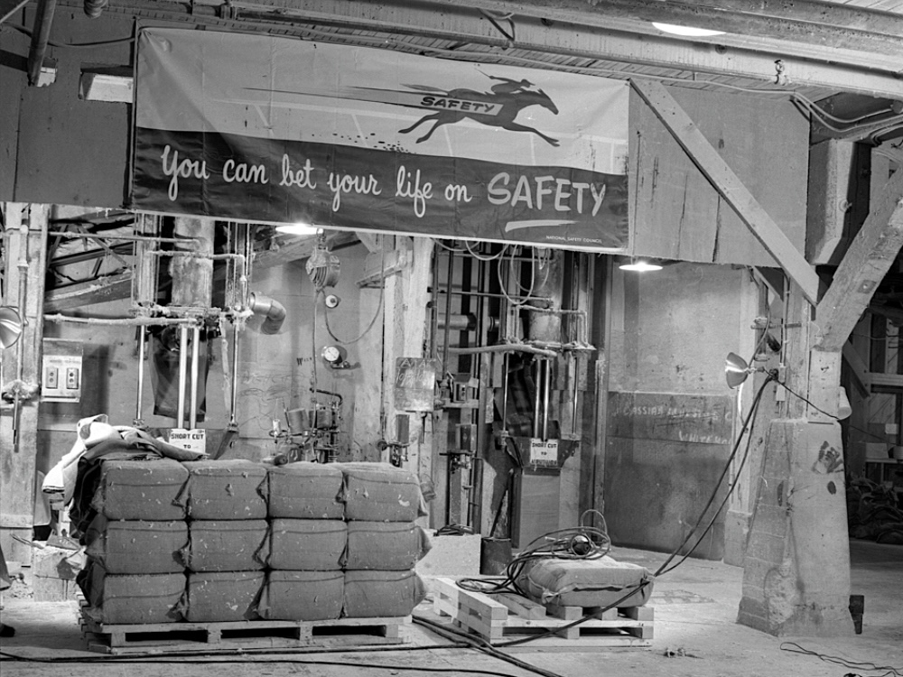 A black and white photo taken inside an industrial-looking space. A banner hanging above stacked bales of asbestos says, &lsquo;You can bet your life on safety.&rsquo;