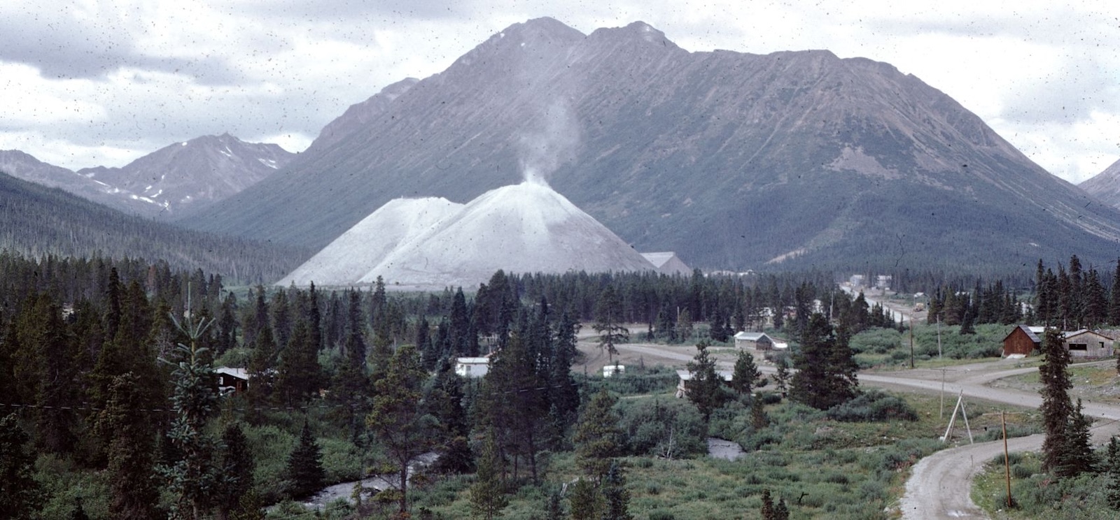 Dust is visible coming from the top of a massive tailings pile looming above a rural community.