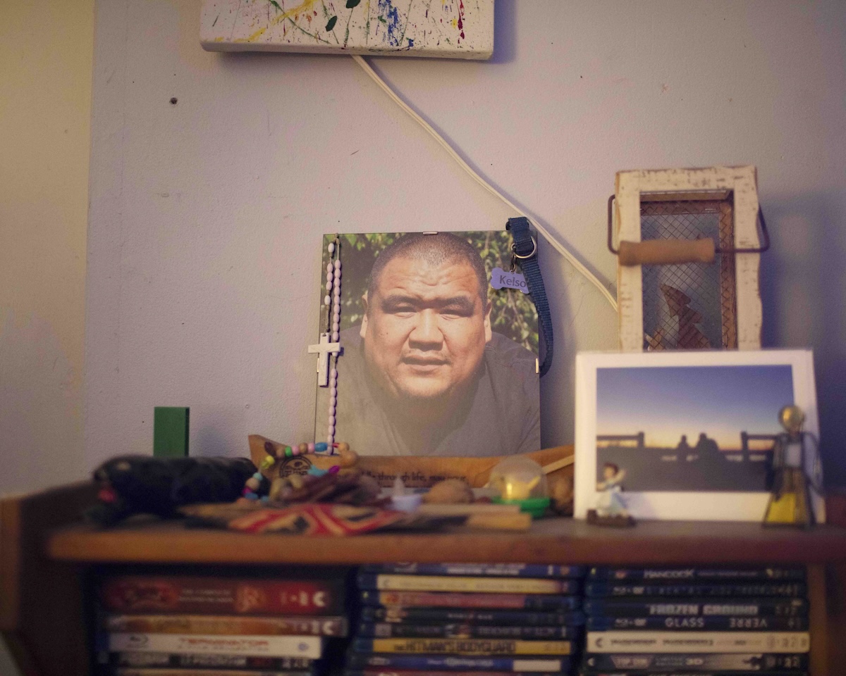 A photo of an Indigenous man named Frankie sits on top of a wooden shelf in a family home. Frankie has short hair in a buzz cut and is wearing a black T-shirt. His photograph is decorated and surrounded with small items meant to honour his memory.