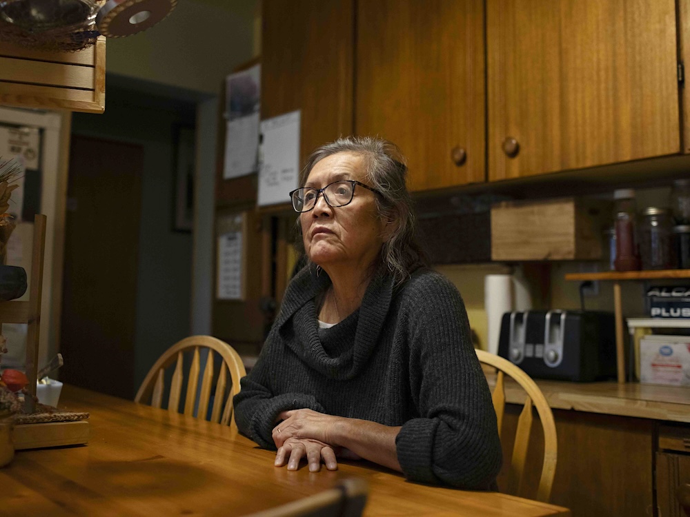 Josephine Andrew is an Indigenous woman with dark greying hair and glasses. She is wearing a black cowl-neck sweater and sitting at her kitchen table with brown wood furnishings.
