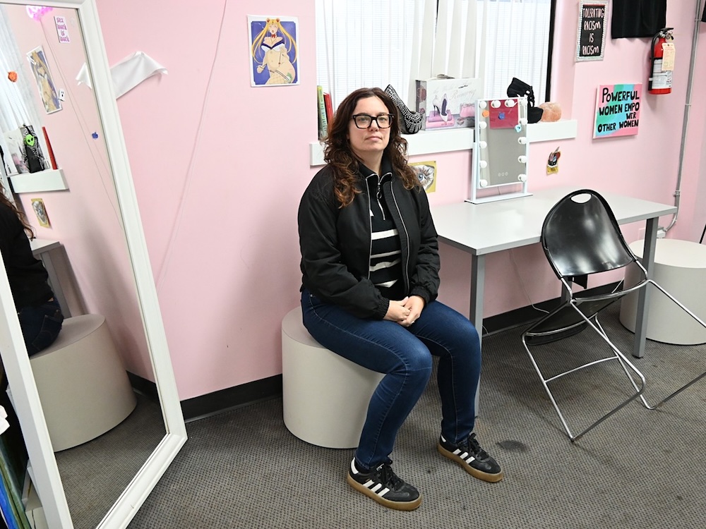 A woman with dark clothes, hair and glasses poses in a pink-walled room.