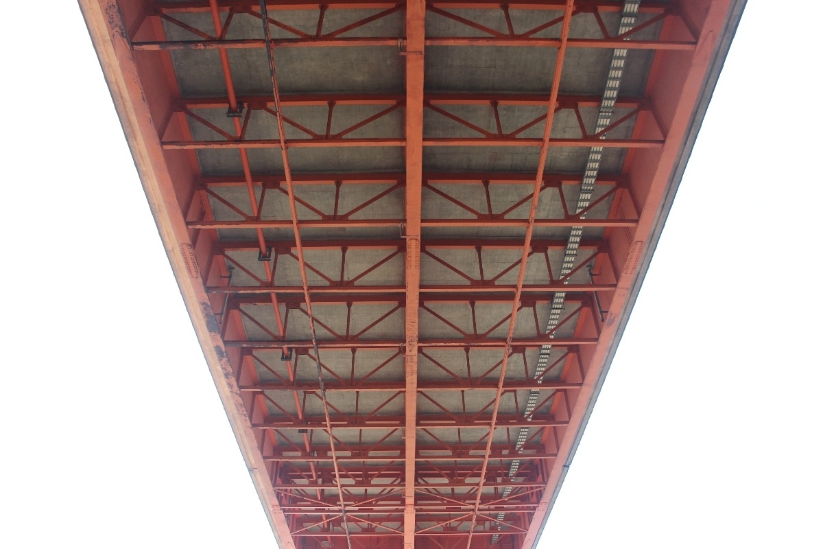 Orange girders in a symmetrical pattern are seen on the underside of a large bridge.