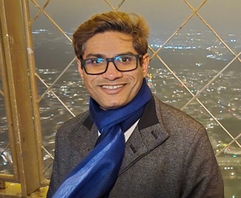 A man, smiling for the camera, wears a blue scarf, blazer and eyeglasses. 