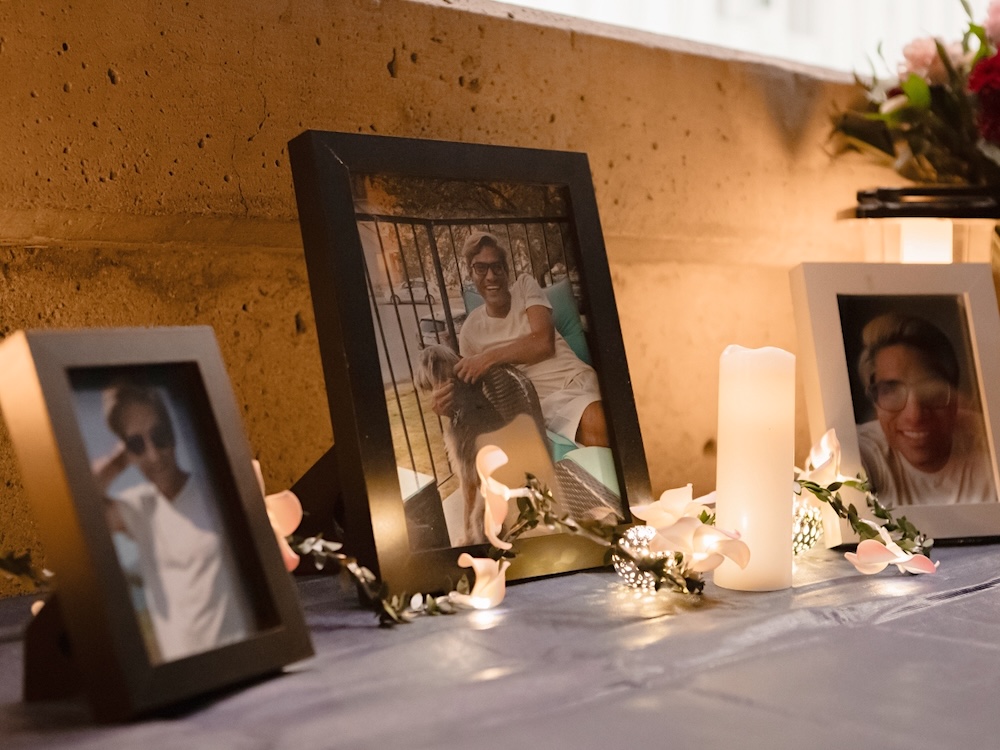 Three framed photos of a smiling man in various poses are displayed on a table with flowers and a candle.