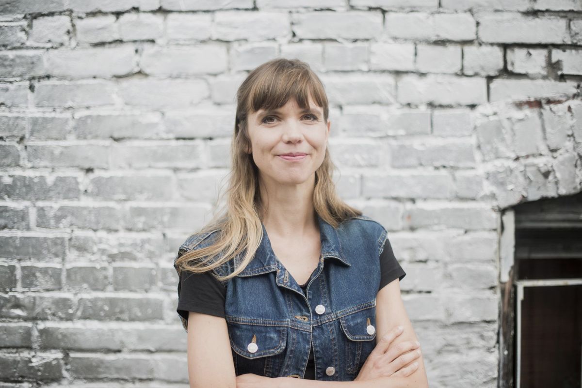 Andrea Kr&uuml;si has long, dirty-blonde hair and a light skin tone. She wears a black T-shirt and denim vest and stands in front of a brick wall.
