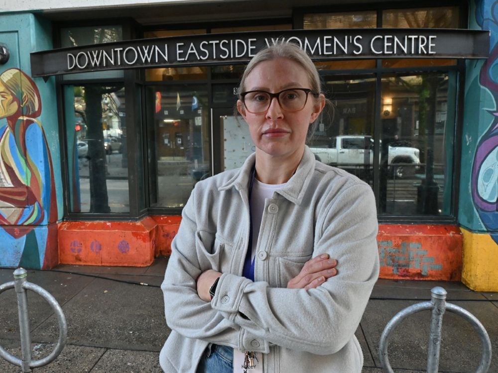 Kaley Merritt has blonde hair that is tied back, and a light skin tone. She is wearing glasses and has her arms crossed. She is standing in front of the Downtown Eastside Women&rsquo;s Centre.