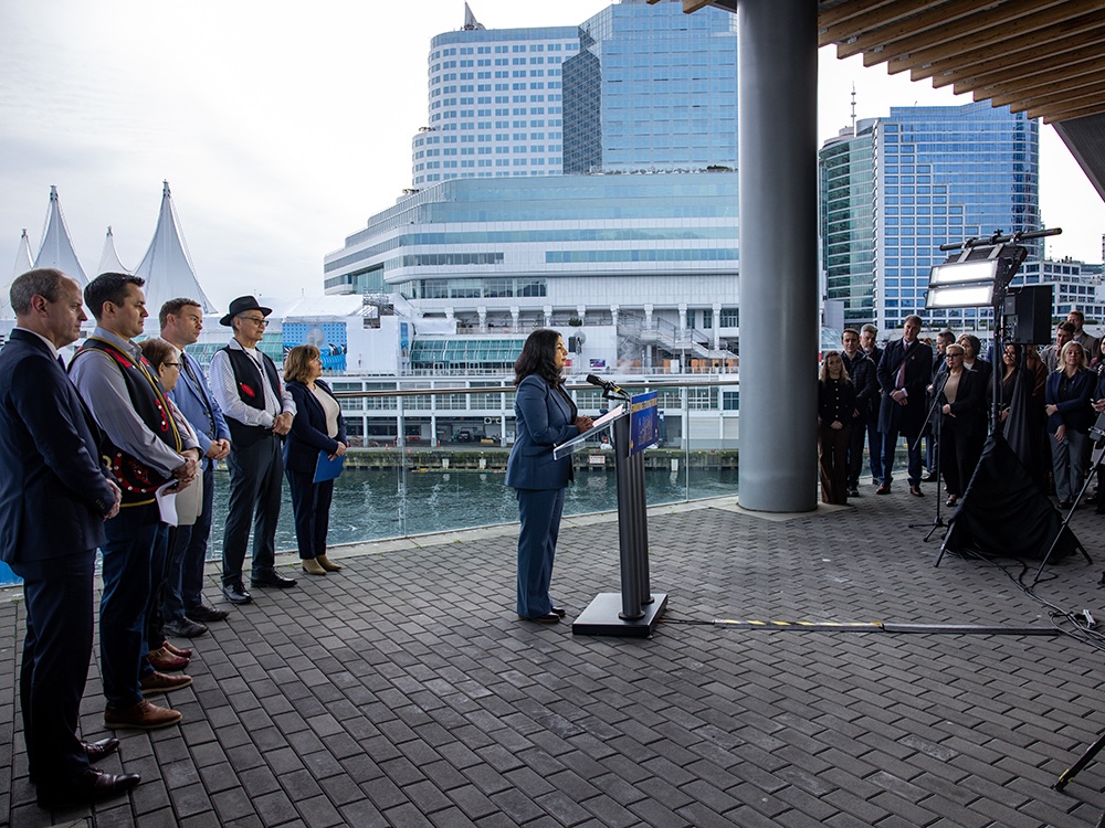 A woman with shoulder-length dark hair wearing a blue pantsuit stands at a lectern with a group of people behind her, viewed from the side. The waterfront Vancouver Convention Centre and Pan Pacific Hotel are in the background.