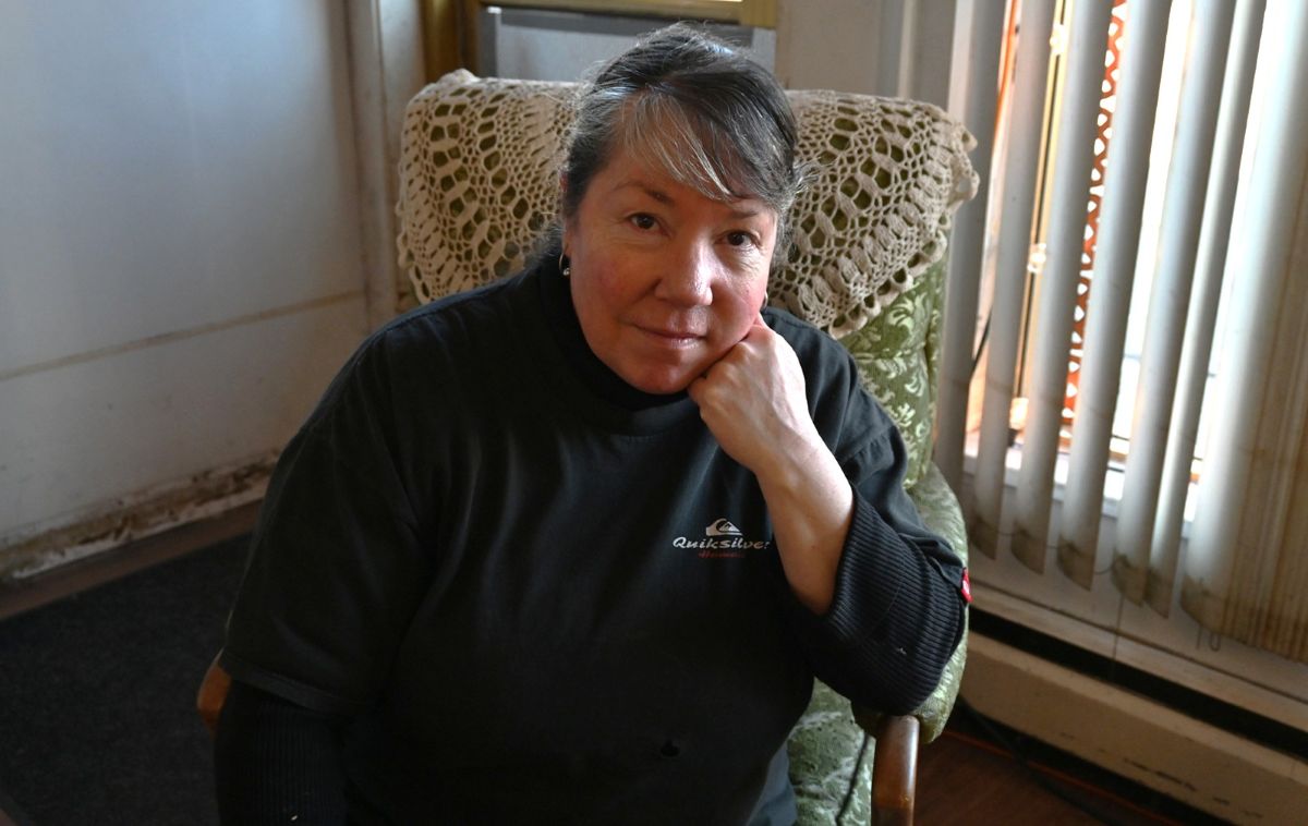 Susan Davis is a middle-aged woman with dark, greying hair and medium-light skin tone. She sits on a green chair covered by a doily, wearing a black Quicksilver shirt.