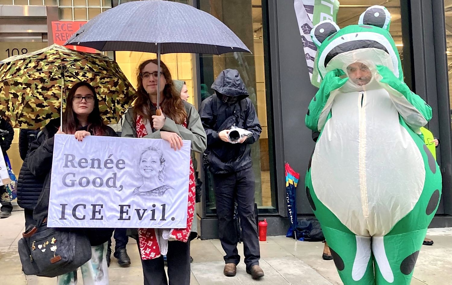 Two protesters hold umbrellas and a sign that reads, &lsquo;Renee Good, ICE Evil.&rsquo; Another protester stands next to them dressed in an inflatable frog costume.