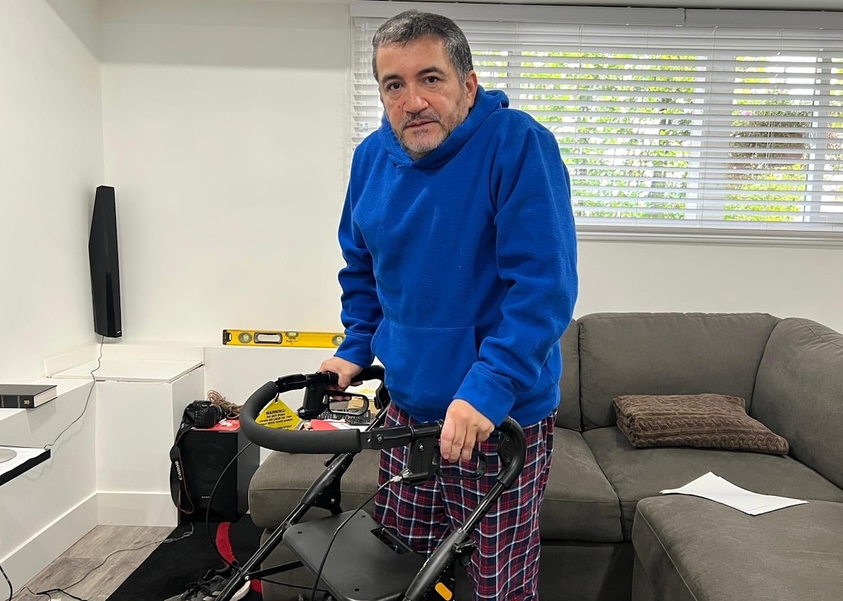 A sick-looking man stands in a sparsely furnished apartment. He is supported by a walker and wears a blue hoodie and pyjama bottoms.