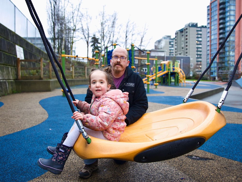 A middle-aged man wearing glasses crouches behind a five-year-old girl who is sitting on a swing at a playground. 