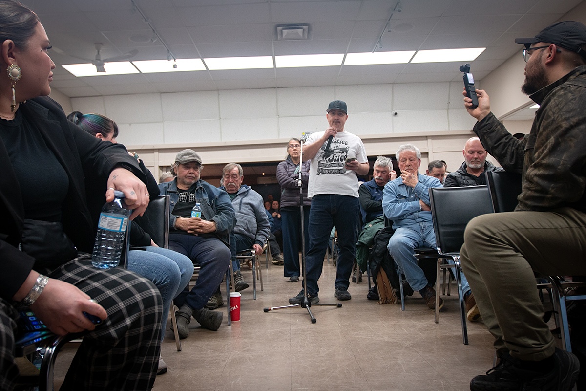 A man in a white shirt and jeans holds a microphone and speaks while flanked by seated attendees.