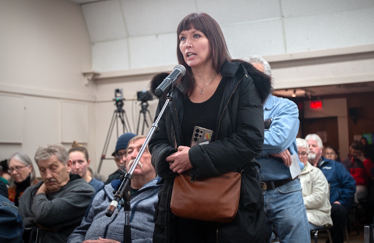A woman in a black coat and wearing a brown bag speaks into a microphone while a seated crowd looks on.