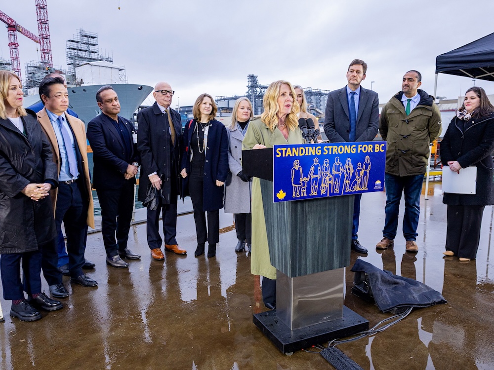 A woman with light skin and long, wavy blond hair speaks at a lectern. Several people stand behind her. Ships can be seen in the background.