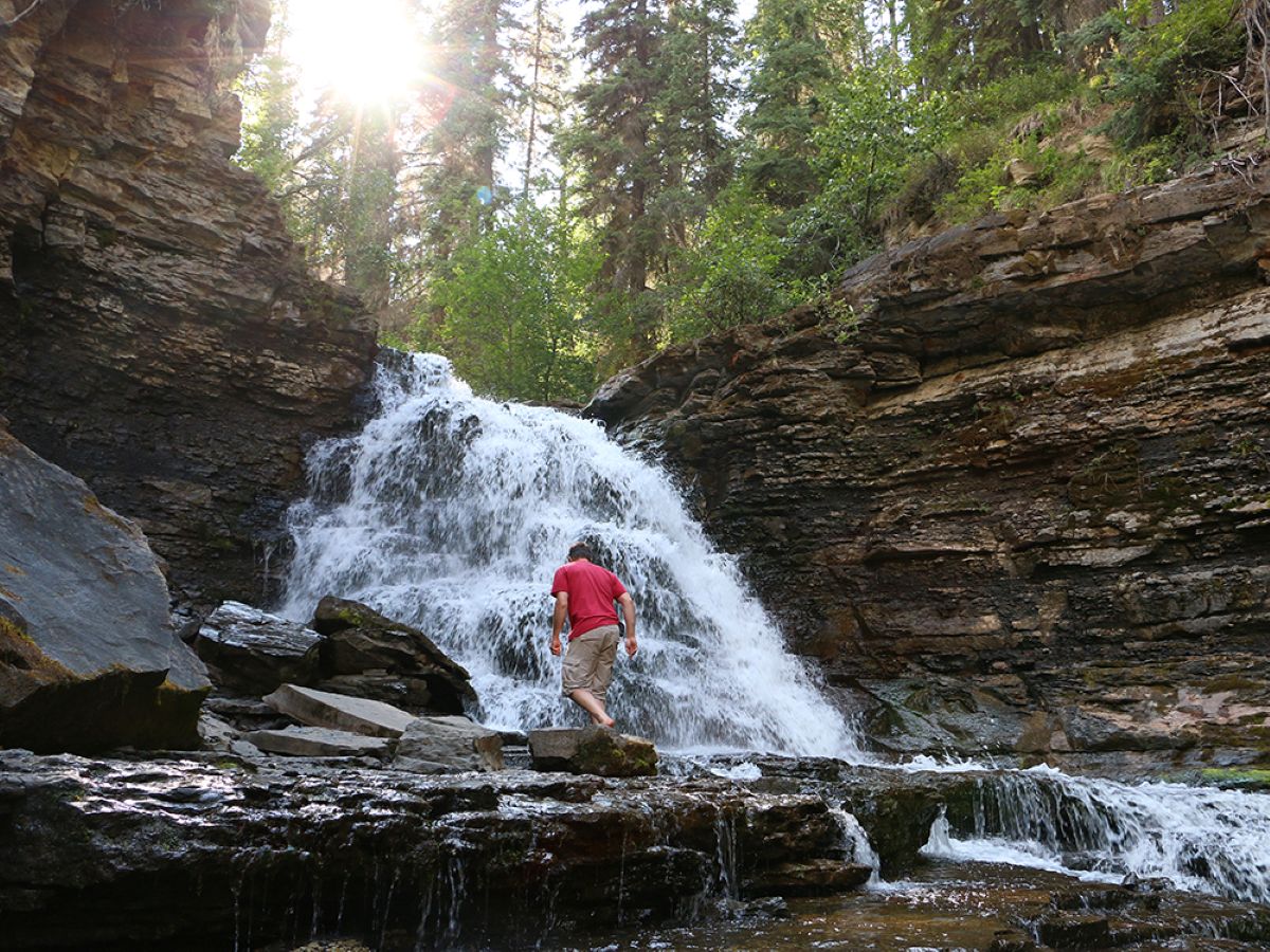 Water cascades over rock in a forest setting as sun peaks through trees. A man walks near the bottom of the falls.