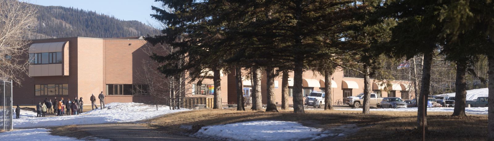 Parents gather in a small group to the left of a two-storey brick school. RCMP trucks are parked on the sidewalk in front of the school entrance. 