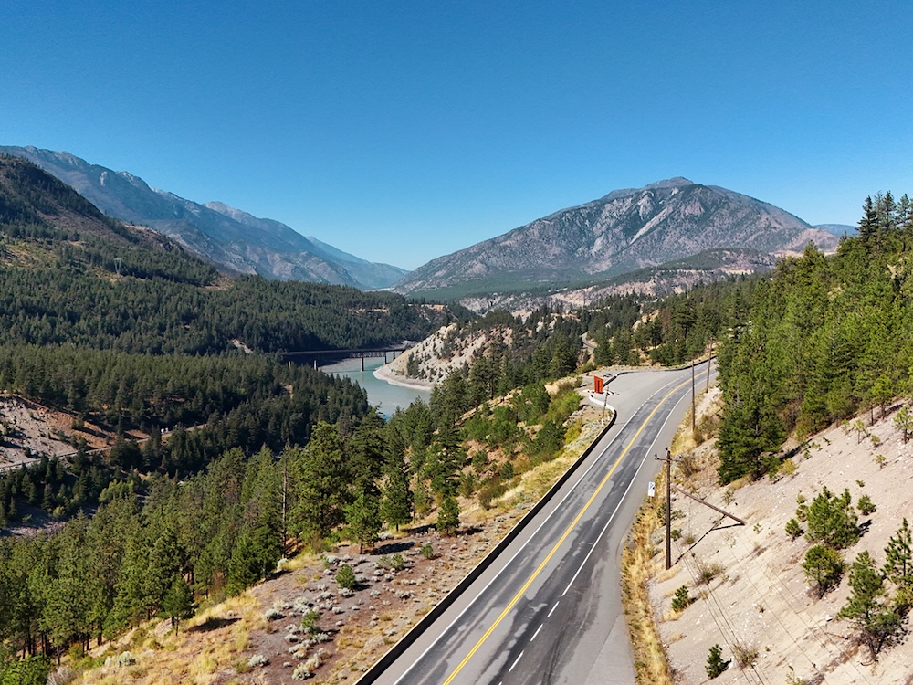 A highway runs through a rugged, sparsely populated valley with steep, forested mountainsides.