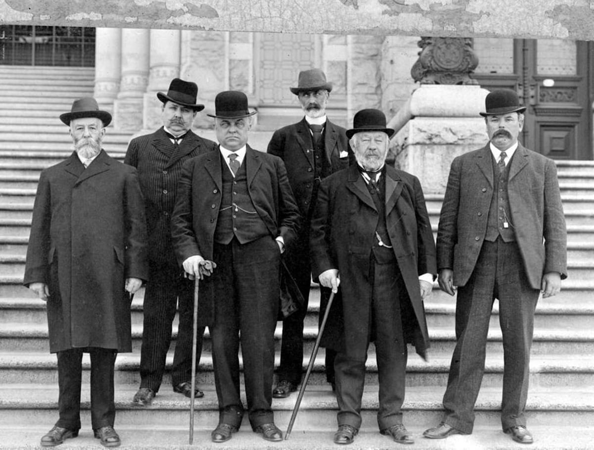 A black and white photo shows six men, all wearing dark clothes and various kinds of formal hats, standing on stone steps.