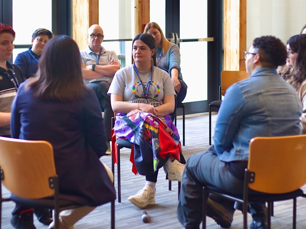 A group of people sit in chairs in a circle inside a sunny conference room at a university.
