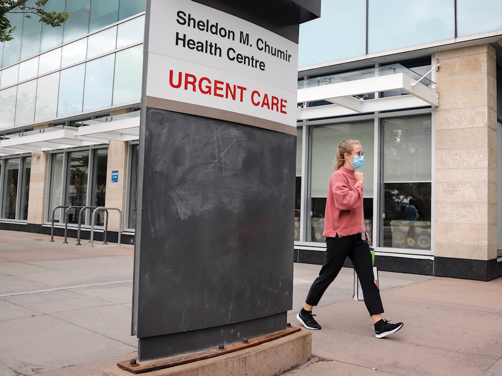 A sign in front of a modern hospital building reads &lsquo;Sheldon M. Chumir Health Centre Urgent Care.&rsquo; A young woman wearing an orange sweatshirt and a mask can be seen walking in front of the building.