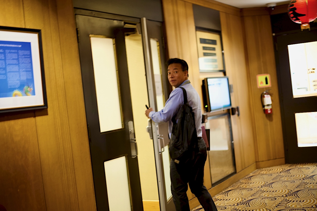 Mayor Ken Sim, wearing a button-down shirt and carrying a shoulder bag, looks over his shoulder as he opens a door at city hall.