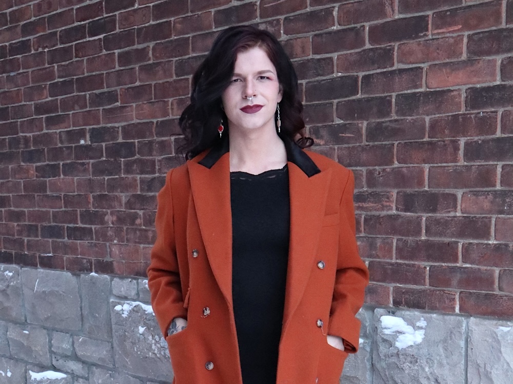 Fae Johnstone has light skin tone and long, wavy black hair. She has a nose ring and is wearing burgundy lipstick. She is wearing a black shirt and an orange wool coat, with her hands in her pockets.