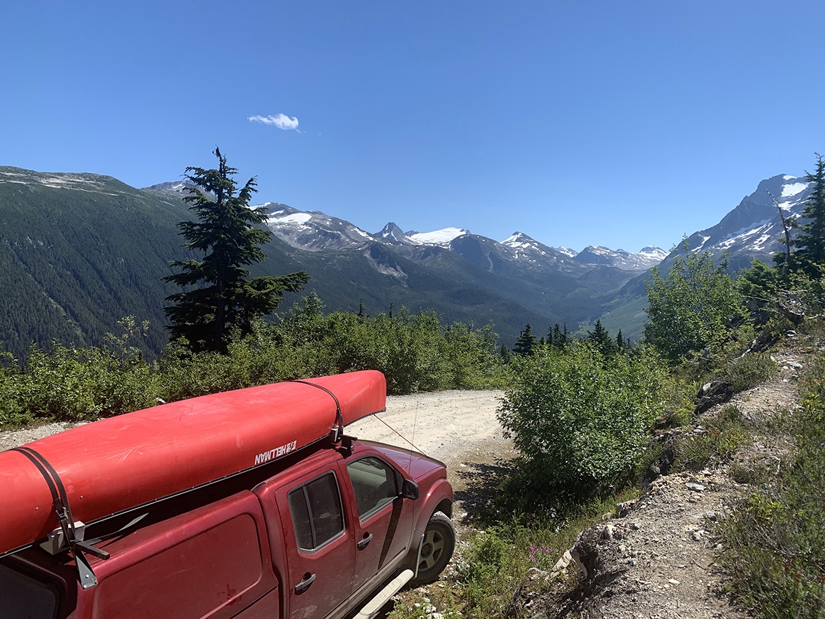 In the foreground is a red pickup truck with a red canoe on the roof. Beyond is a mountain landscape and blue sky.