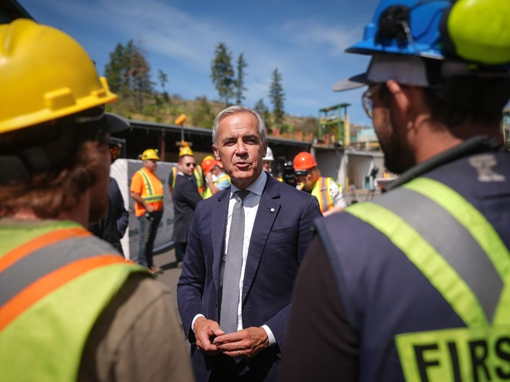 A man in his 60s with light skin and grey hair and wearing a suit speaks to two men who stand with their backs to the camera, wearing hard hats and safety vests.