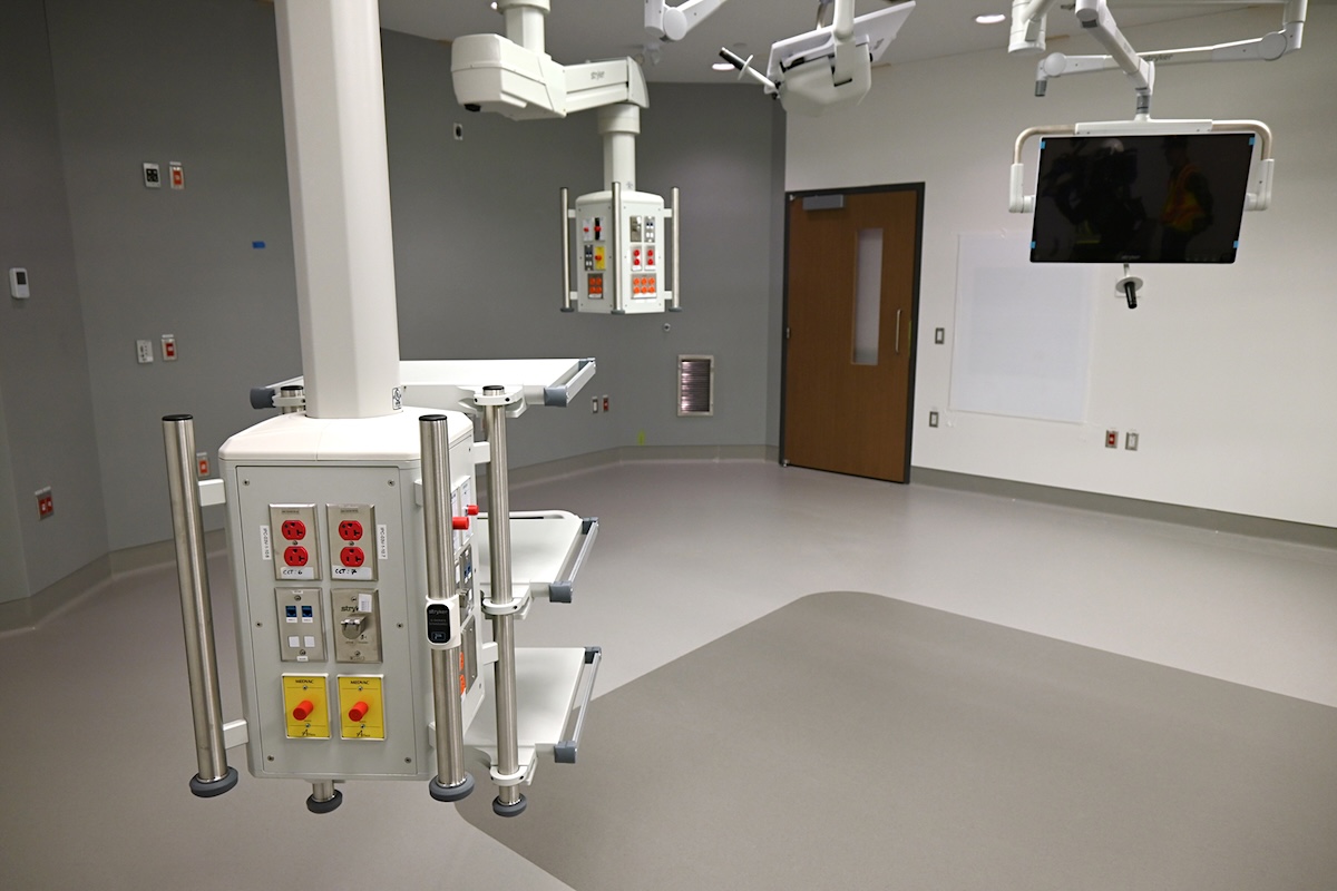 A large white room has various boxes hanging from the ceiling on mechanical arms. Tables, electrical plugs and brightly coloured switches and knobs are on the side of one of the boxes closest to the camera. 