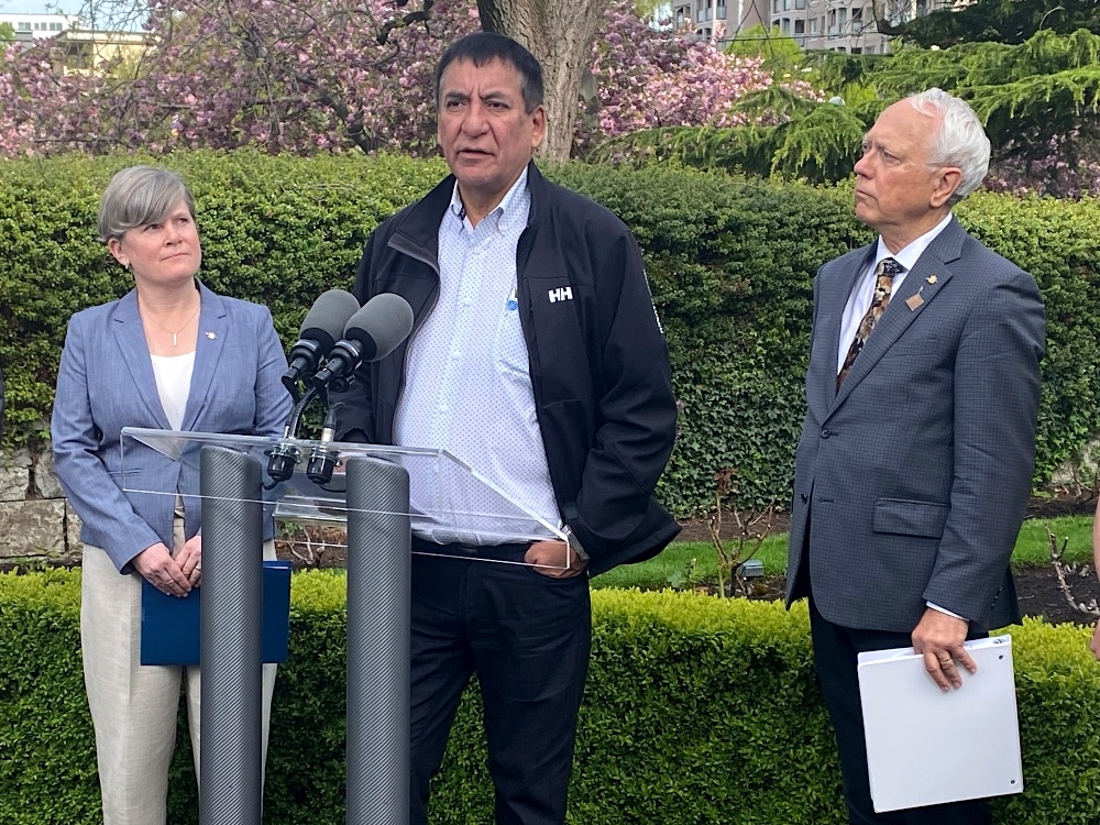A tall Indigenous man with dark hair speaks into a microphone, flanked by two grey-haired people with fair skin.