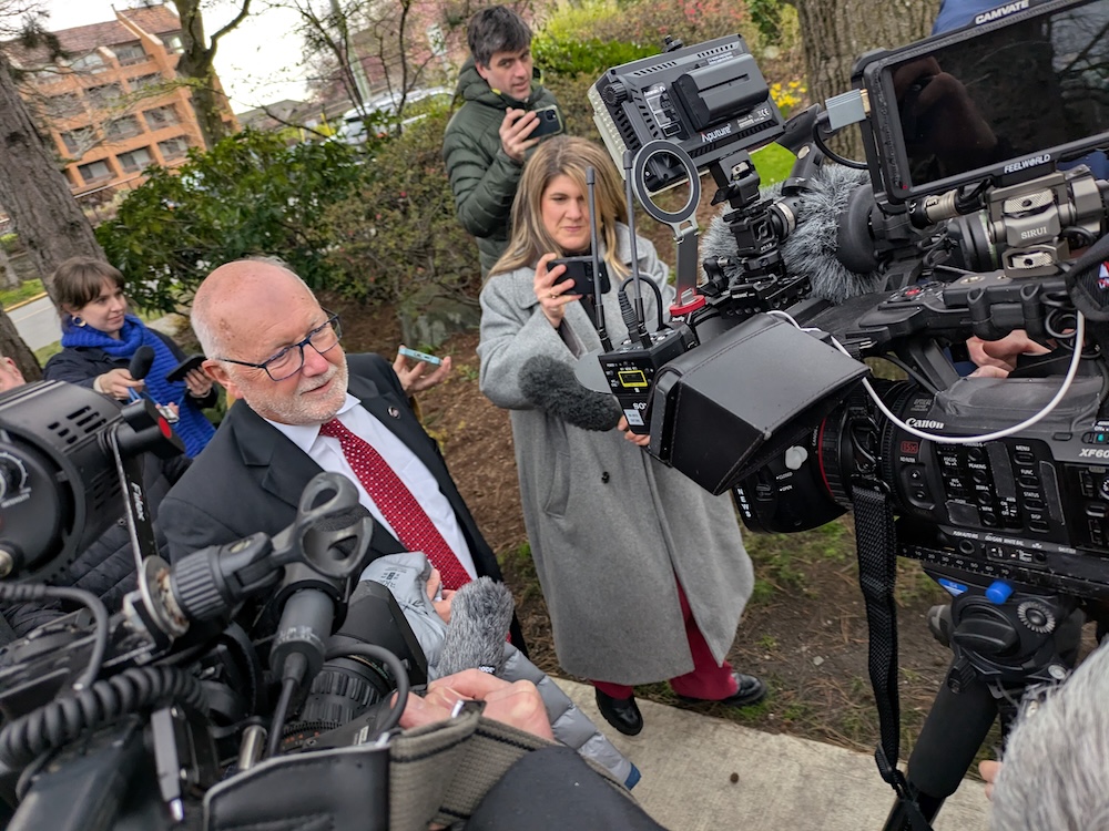An older man with a trimmed white beard is surrounded by media cameras.