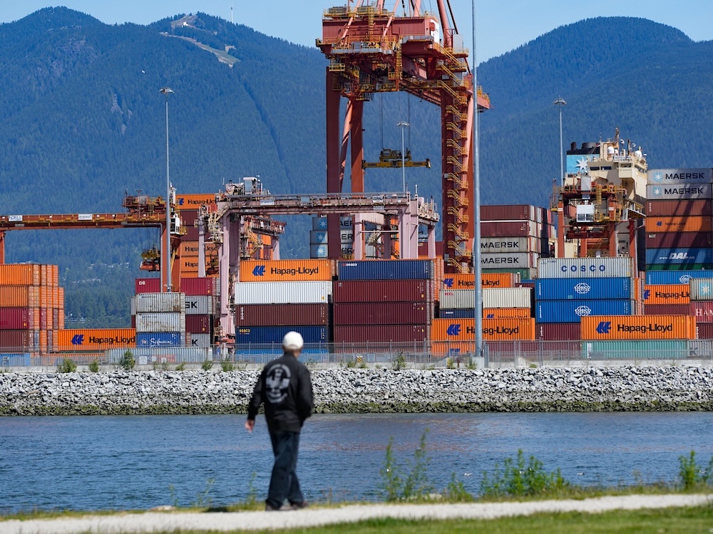 Orange, brown, blue and white shipping containers stacked near and under a gantry crane at one of Vancouver&rsquo;s port terminals. The North Shore mountains can be seen in the background.