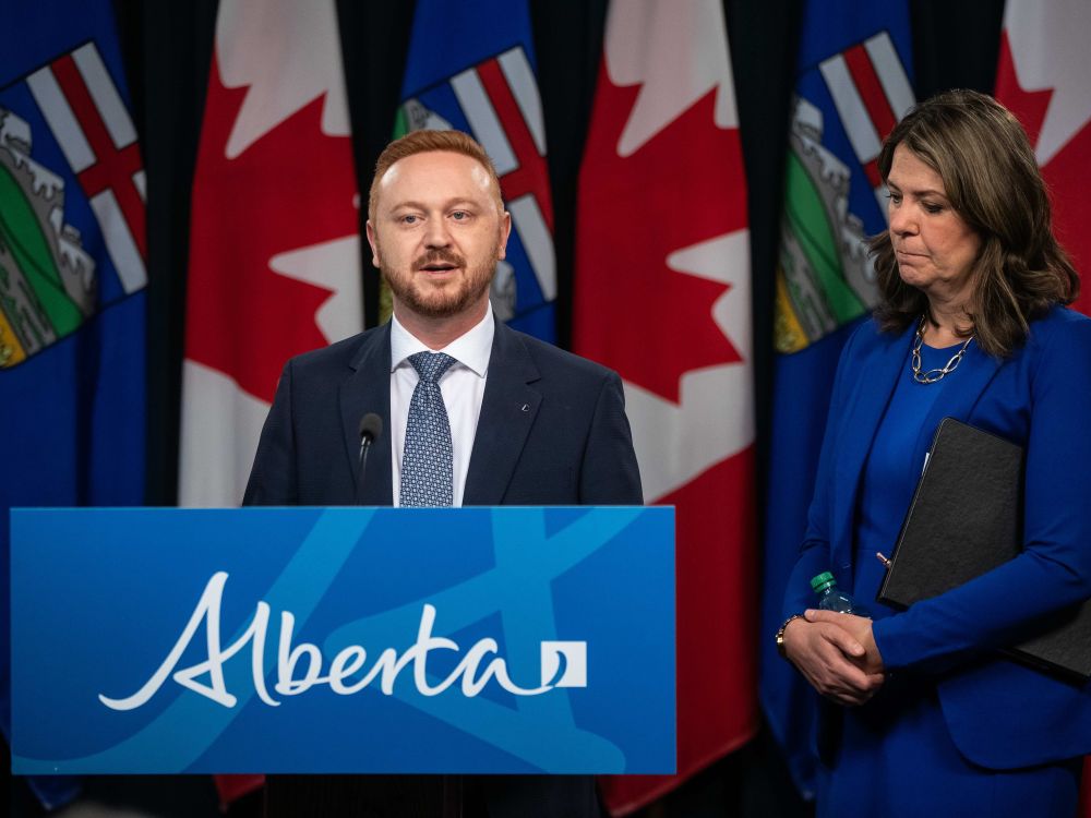 A 40-ish light-skinned man with red hair stands at a podium as Alberta Premier Danielle Smith looks on.